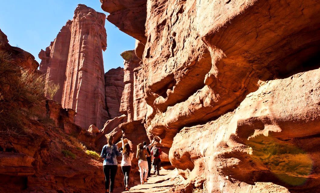 A group of people hike along a narrow trail surrounded by the tall, rugged Velasco red rock formations under a clear blue sky.