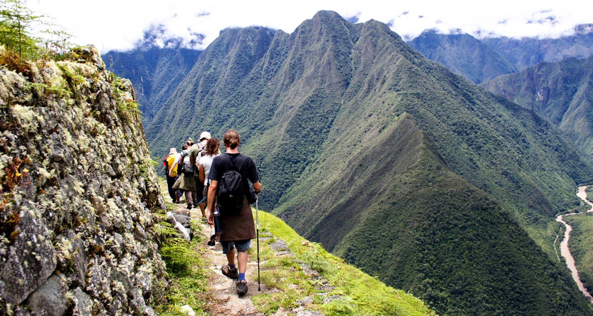 A group of hikers walk along the narrow Inca Trail, surrounded by lush green peaks, with a river visible far below in the valley on their way to Machu Picchu.