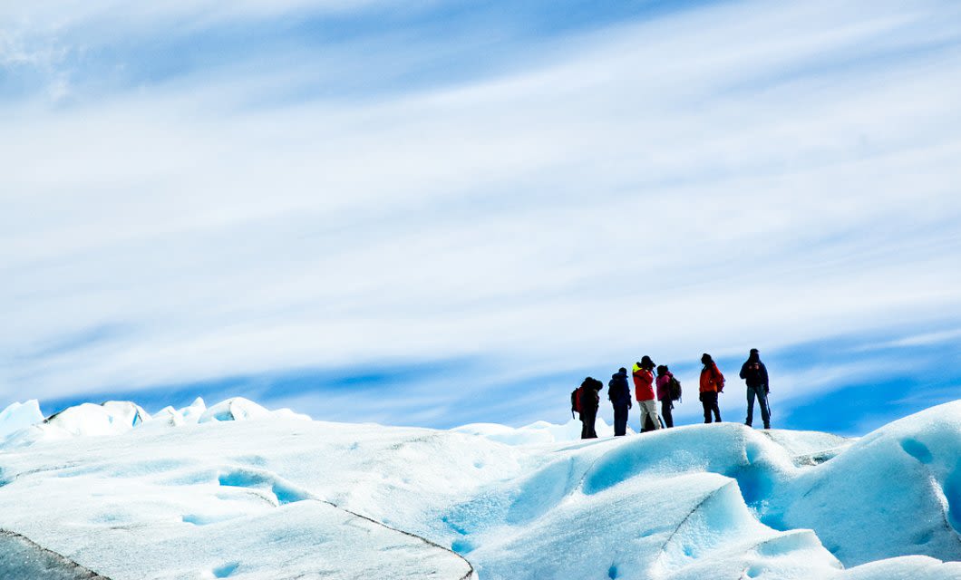 A group of people wearing winter clothing stand on a snowy, icy Patagonia landscape under a blue sky with wispy clouds.