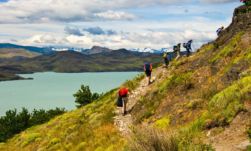 A group of hikers with backpacks walks up a rocky trail on a green hillside overlooking a blue lake in Tierra del Fuego, with distant mountains and a partly cloudy sky in the background.