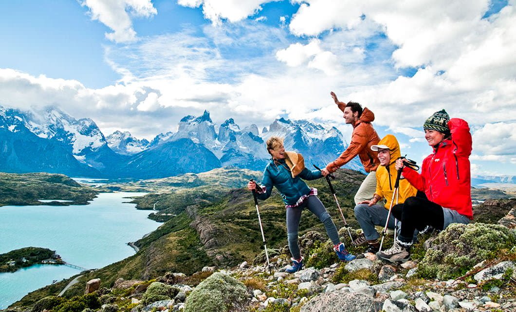 Four hikers in colorful jackets smile and pose with trekking poles on a rocky hilltop, overlooking the scenic Torres del Paine landscape—majestic mountains, dramatic clouds, and a winding turquoise river below—a perfect moment from their unforgettable tour.