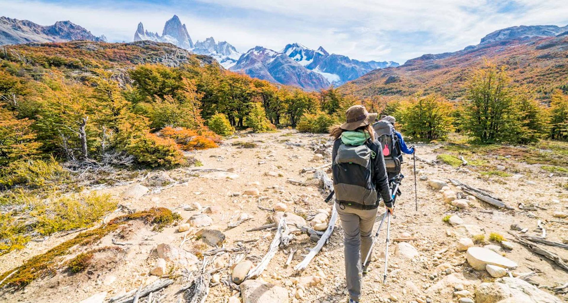 Two hikers with backpacks and trekking poles walk along a rocky trail surrounded by autumn trees near El Chaltén, with snow-capped mountains and dramatic peaks in the background under a partly cloudy sky, perfect for a trekking tour.