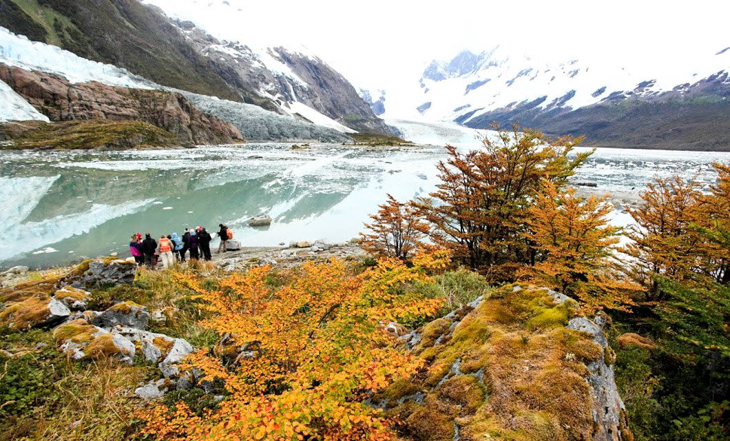A group of people stands near the shore of a glacial lake in Patagonia, surrounded by mountains and autumn-colored foliage, with icebergs floating on the water and snow-capped peaks in the background.