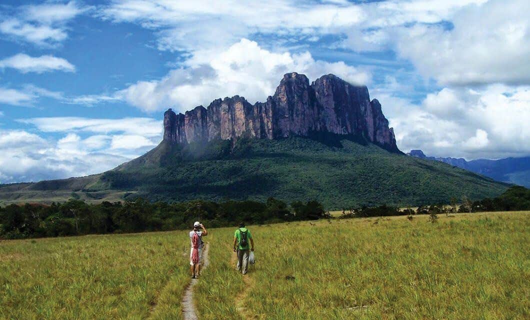 Two hikers walk on a grassy trail toward Mount Roraima’s flat-topped summit with steep cliffs under a partly cloudy sky. Dense green trees surround the base, capturing the adventure of a classic Mount Roraima Trek.