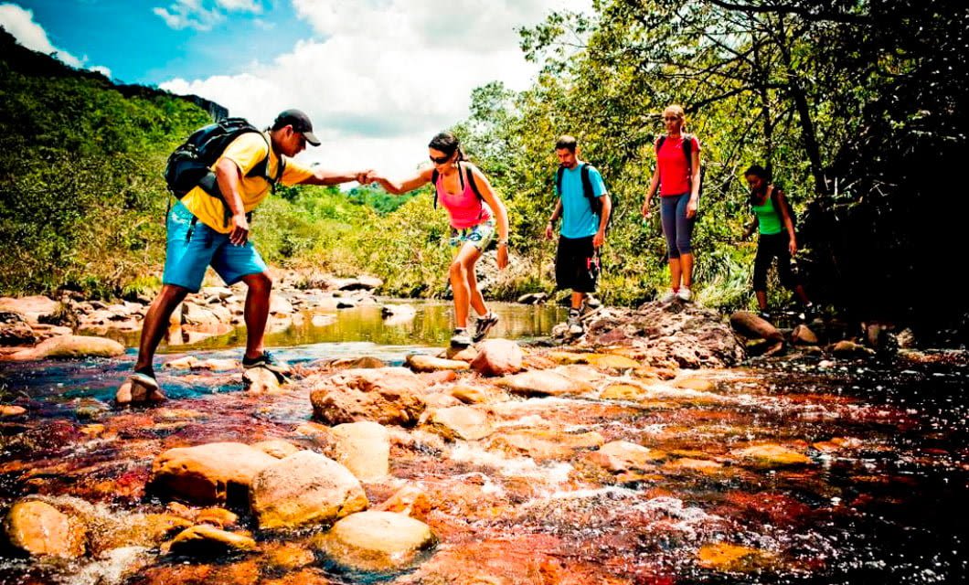 A group of five hikers crosses a shallow rocky stream in a lush Santa Catarina outdoor setting. One person helps another step over the water, while others wait behind, all wearing casual hiking gear and backpacks on their Brazil Adventure Tour.