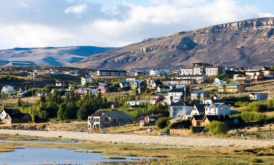 A scenic view of a small town, reminiscent of El Chalten, with colorful houses nestled at the base of rocky hills, a river and grassy banks in the foreground—an inviting spot for any trekking tour under a partly cloudy sky.