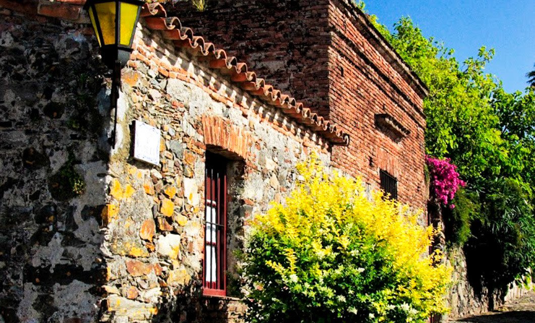 A rustic stone and brick building with a tiled roof stands beside a bright yellow-green bush and magenta flowers, under a clear blue sky and vintage streetlamp—capturing the timeless charm found on Estancias of Uruguay.