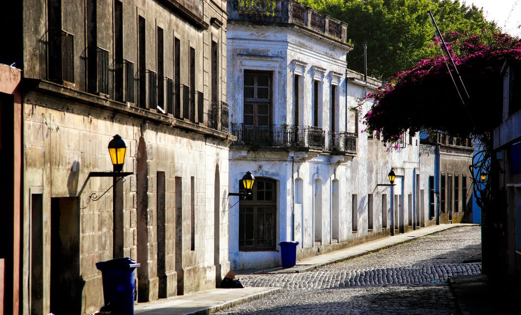 A quiet cobblestone street lined with old, weathered buildings, black iron balconies, and yellow-lit lanterns; purple flowers hang over the sidewalk and blue trash bins sit along the Coast to Coast Tour route.