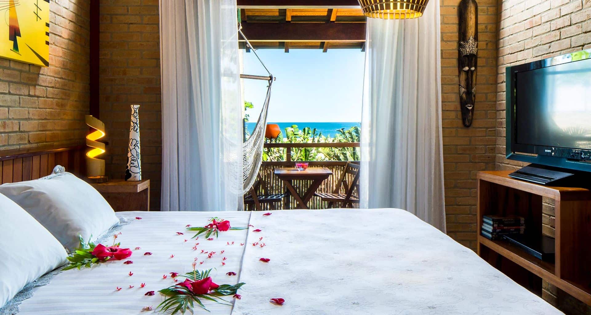 A cozy bedroom with a bed decorated with pink flower petals, a TV, and a small balcony. The balcony has a hammock, chairs, and table—perfect for relaxing after a luxury adventure tour of Lencois Maranhenses under the bright blue ocean sky.