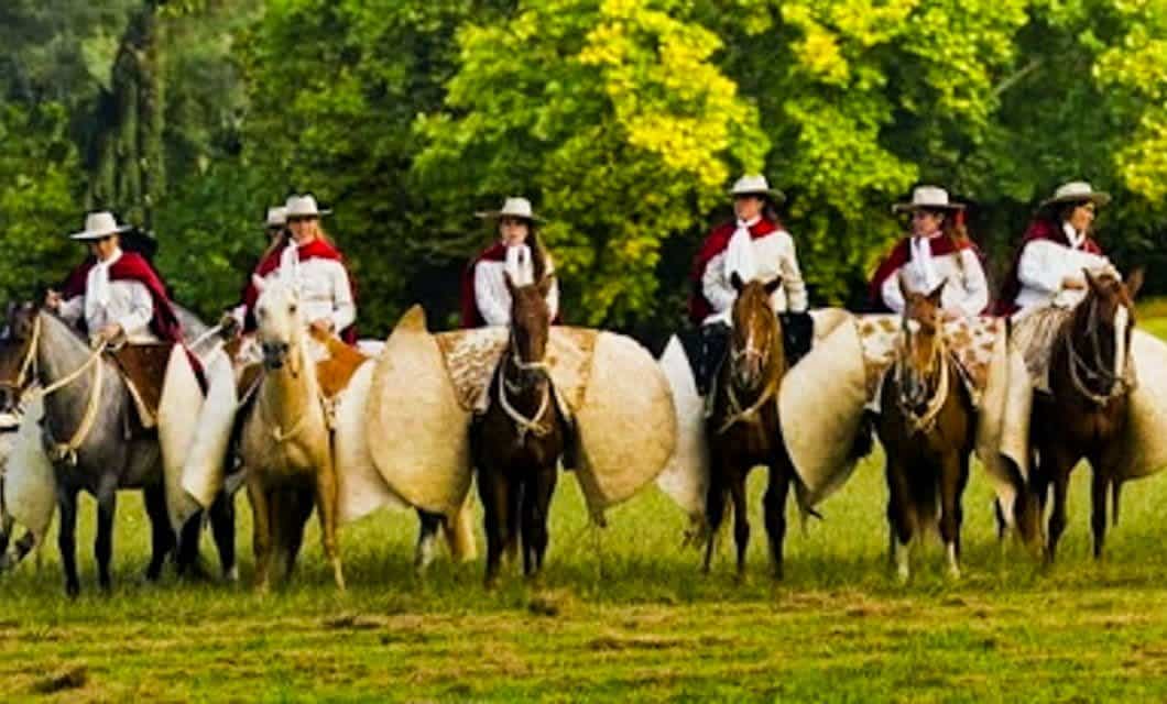 A group of people in white shirts, red capes, and hats sit on horses lined up outdoors along the Salta Wine Route, each horse decorated with large round white adornments; trees create a green background.