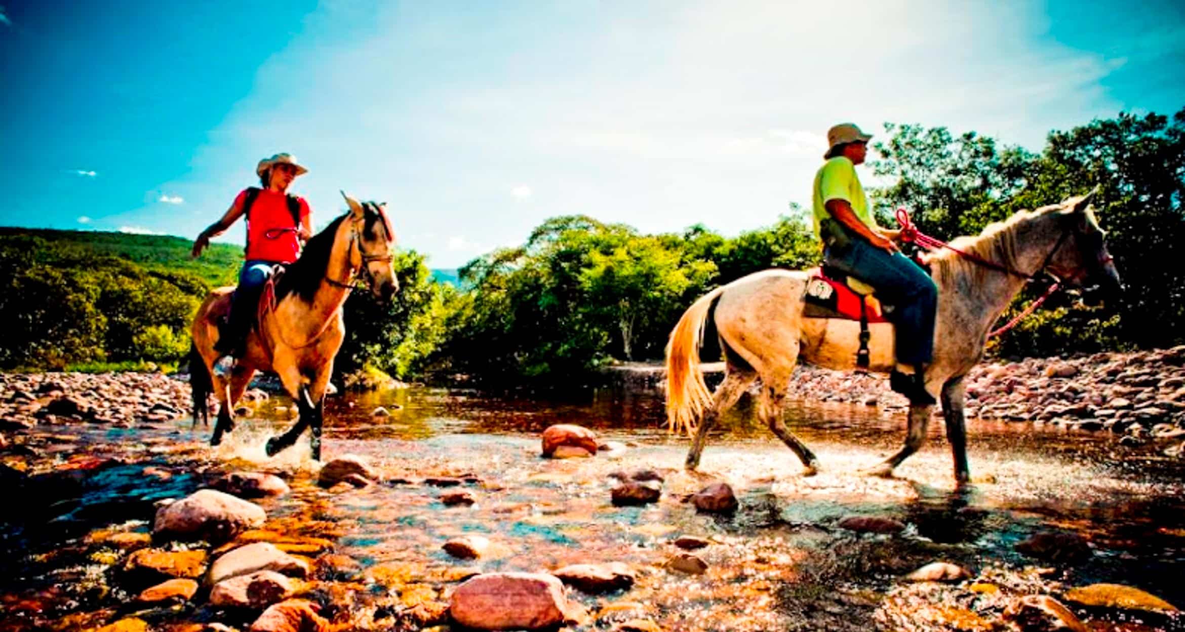 Two people enjoy a horseback riding adventure through a shallow, rocky stream surrounded by lush trees and greenery in sunny Southern Brazil. One rider wears a hat and orange shirt, the other a green shirt and cap.