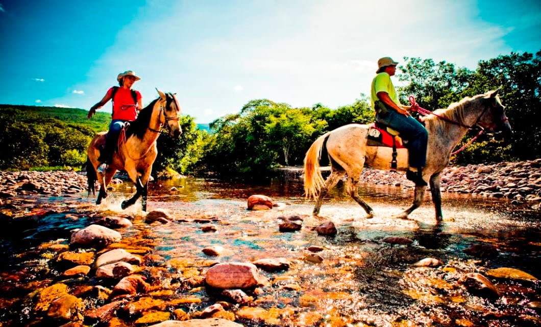 Two people enjoy an adventure horseback riding through a shallow rocky stream under a bright blue sky in southern Brazil, surrounded by lush green trees and distant hills as sunlight reflects off the water and stones.