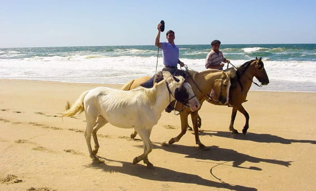 Two people riding horses along a sandy beach near Piriapolis, near the ocean. One person waves a hat while a third horse walks alongside them. The sea and sky are visible in the background—a perfect scene for your next vacation package.