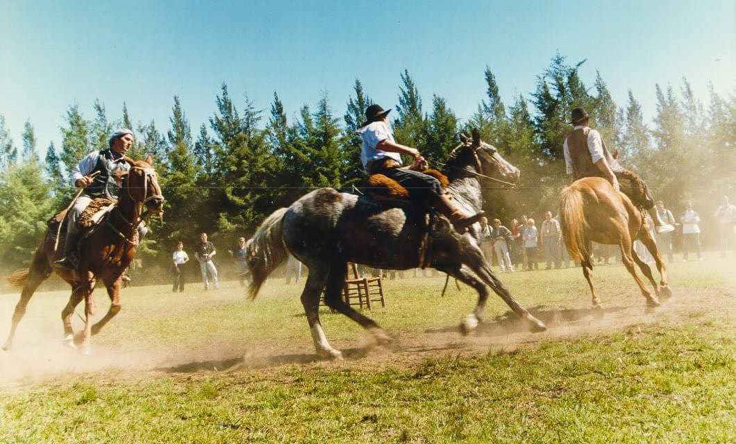 Three people in traditional clothing ride horses quickly across a grassy field during a Paraguay Tour, kicking up dust as spectators watch near tall trees under a clear sky.