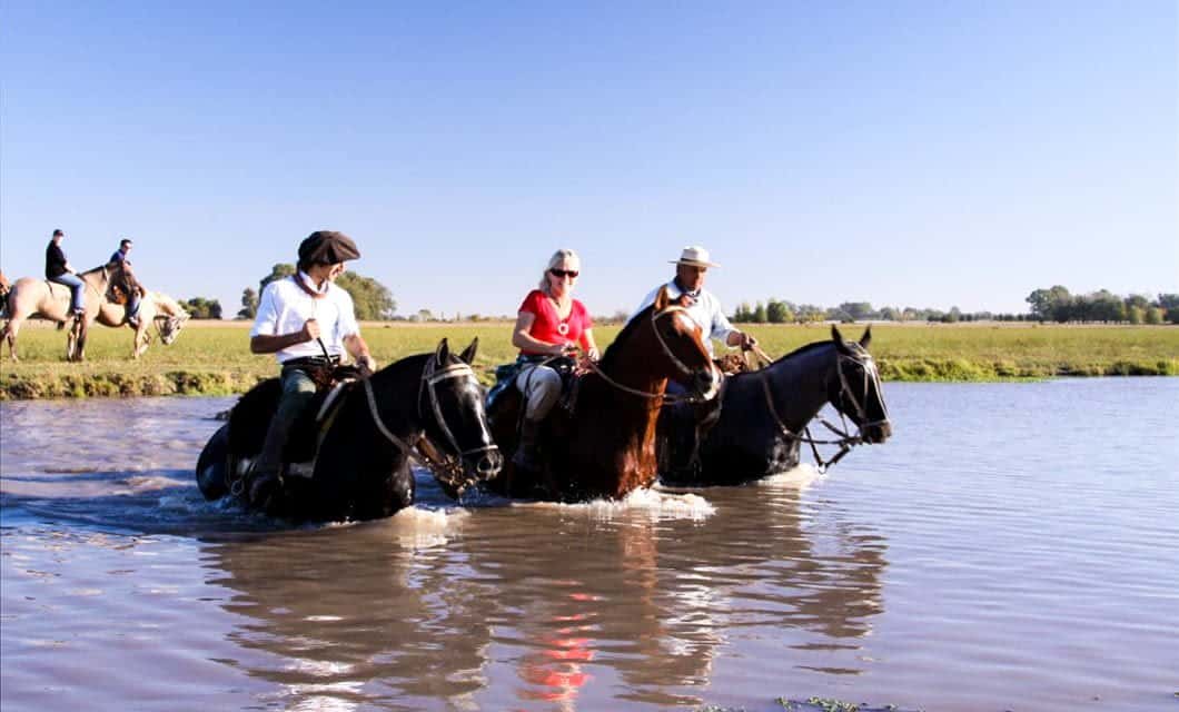 Three people riding horses cross a shallow river in Uruguay, with grassy fields and other riders in the background under a clear blue sky.