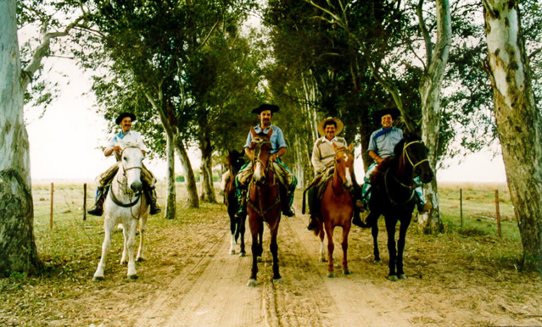 Four people enjoy horseback riding down a dirt path lined with tall trees in rural Uruguay, wearing traditional clothing and hats. The relaxed scene captures the country’s natural beauty and heritage.