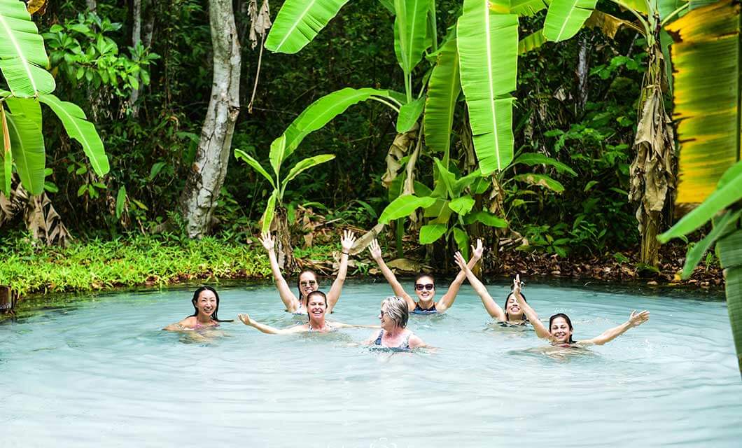 A group of seven people smile and raise their arms while swimming in a natural, blue-tinted pool surrounded by lush green tropical plants and banana leaves on a tour of Brazil's Cerrado in stunning Jalapão.