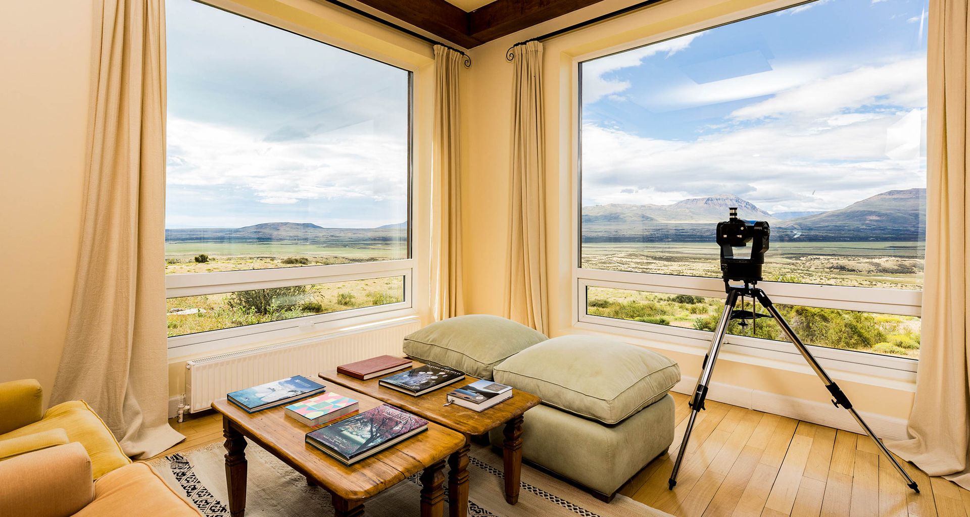 A cozy room at Eolo Patagonia Lodge features large windows with scenic views of Patagonia’s mountains; a telescope faces the window, while a table with books rests between two armchairs and a soft ottoman in this luxury lodge.