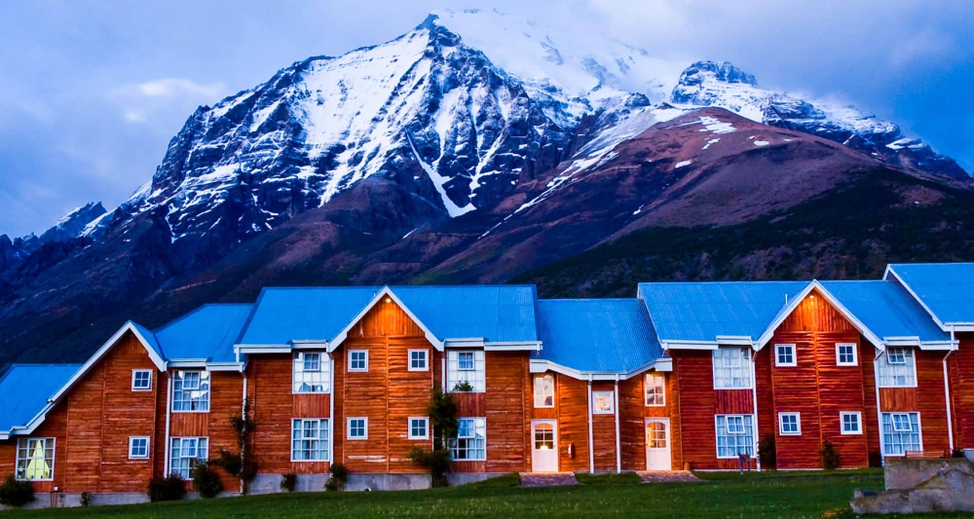 A row of wooden lodges with blue roofs sits at the base of a dramatic, snow-capped mountain under a cloudy sky, warm lights glowing from the windows—welcoming guests to Hotel Las Torres.