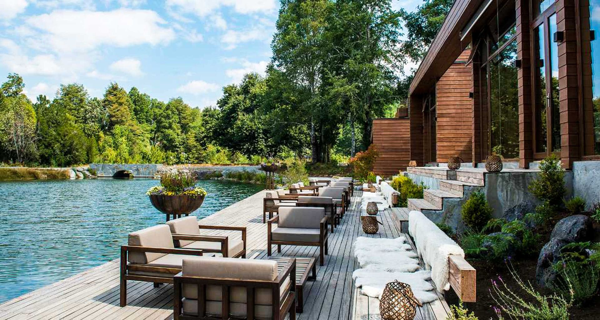 A wooden deck with cushioned chairs and tables overlooks a serene lake surrounded by trees at Hotel Vira Vira. The deck, attached to a modern wooden building, features stairs and decorative planters for a cozy, inviting atmosphere.