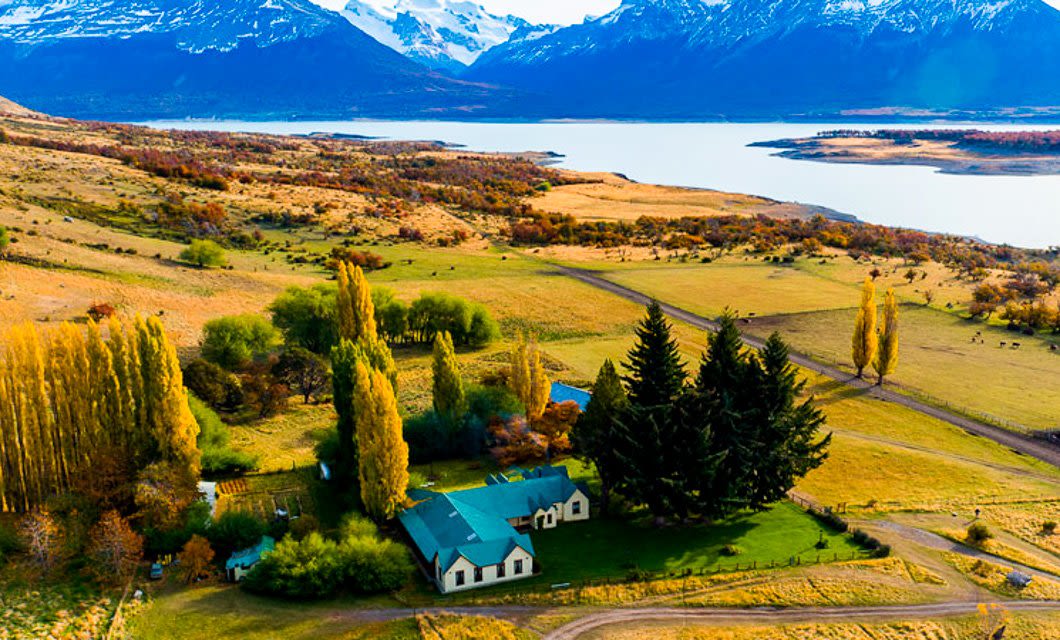 A cluster of white houses with green roofs sits among trees on a grassy landscape, with a lake and snow-capped mountains in the background under a clear sky—perfect for an El Calafate or Patagonia Eco Tour experience.