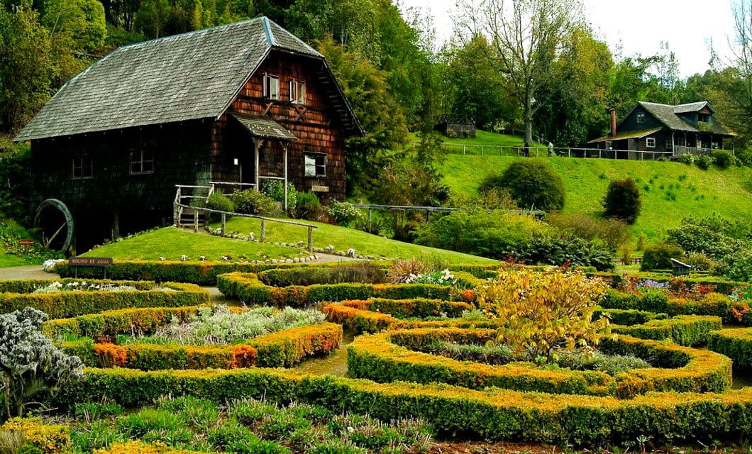 A rustic wooden house with a water wheel sits on a grassy hillside in Patagonia, behind a colorful, neatly trimmed flower garden with winding hedges. Trees and another house are visible in the background.