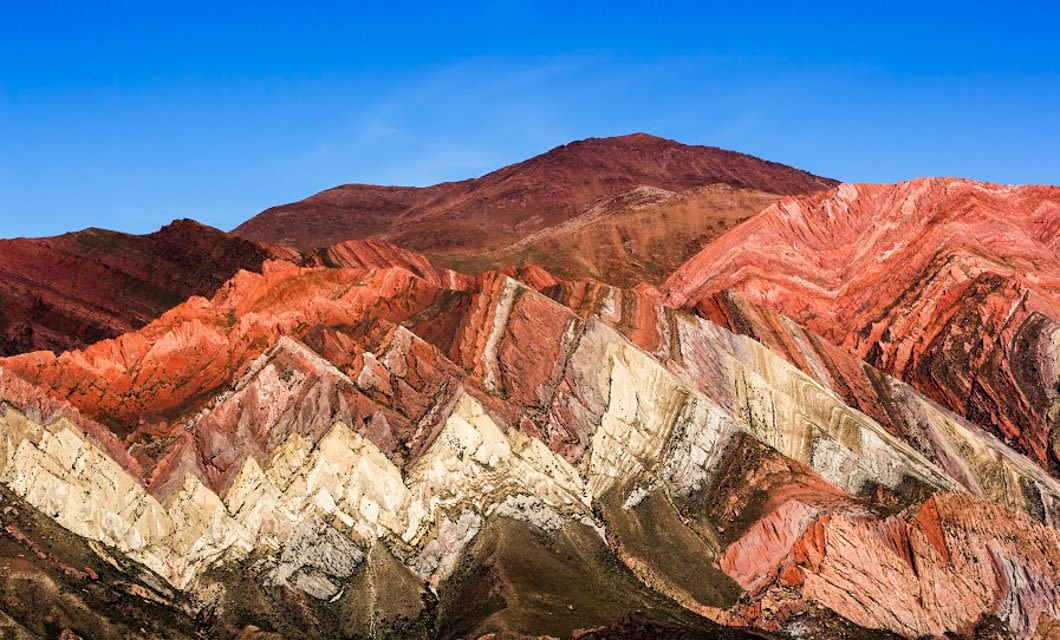 Colorful, jagged mountains in Argentina’s Land of Contrast reveal layers of red, pink, white, and beige rock formations under a clear blue sky, creating a striking and vibrant landscape.