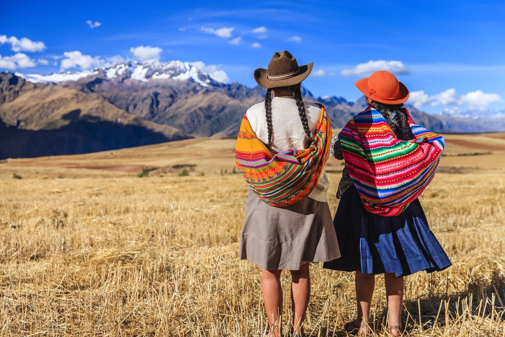 Two women in traditional Andean clothing and hats stand in a golden field, facing distant snow-capped mountains—a breathtaking scene that captures the natural wonders of South America under a bright blue sky with scattered clouds.