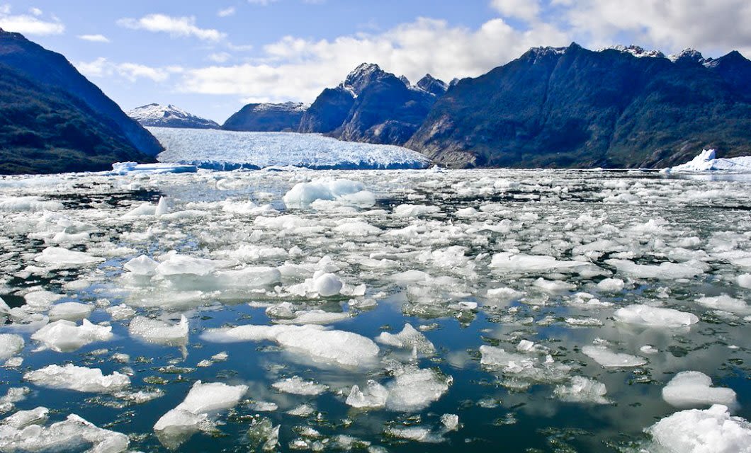 Floating icebergs and chunks of ice cover a cold, reflective body of water, with a glacier and snow-capped mountains in the background—an iconic scene on a Patagonia Glaciers Cruise.