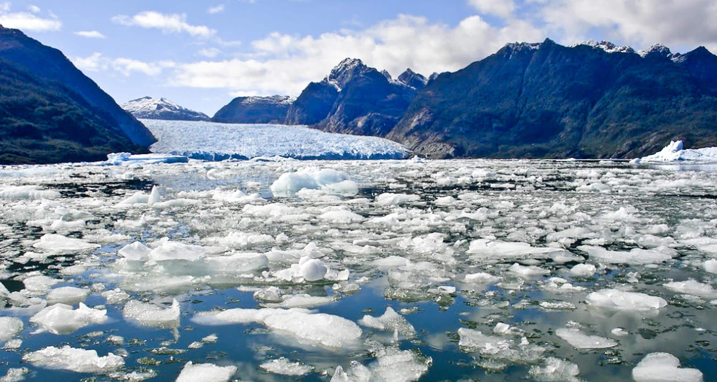 Floating ice chunks cover a cold body of water, with a large glacier and rugged, snow-dusted mountains in the background—capturing the breathtaking scenery of a Patagonia Glaciers Cruise under a partly cloudy sky.