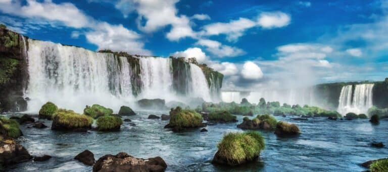 Wide view of a large, powerful waterfall cascading over cliffs, with mist rising and lush green vegetation covering scattered rocks in the flowing river under a partly cloudy blue sky.