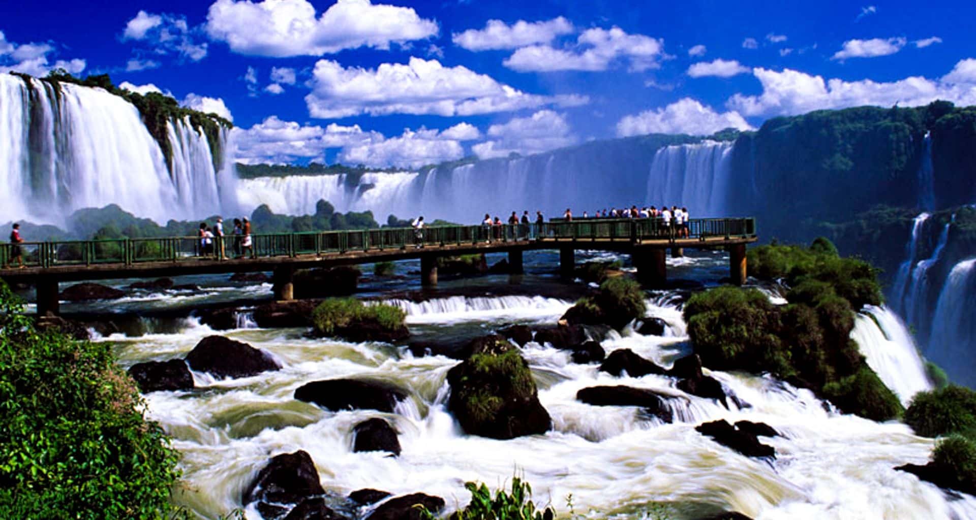 A group of people stands on a wooden bridge overlooking the powerful Iguazu Falls, surrounded by lush greenery, under a bright blue sky with scattered clouds—a breathtaking sight on their Paraguay tour.