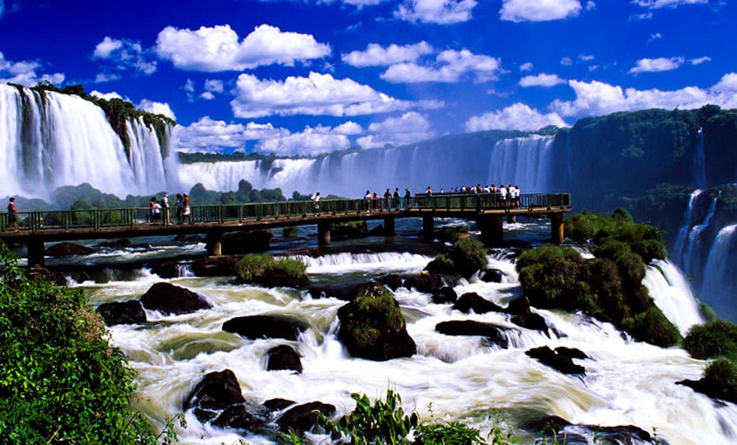 A viewing platform extends over rushing water and rocks, with people on tour observing the dramatic Iguazu Falls, surrounded by lush greenery near Paraguay under a bright blue sky with scattered clouds.