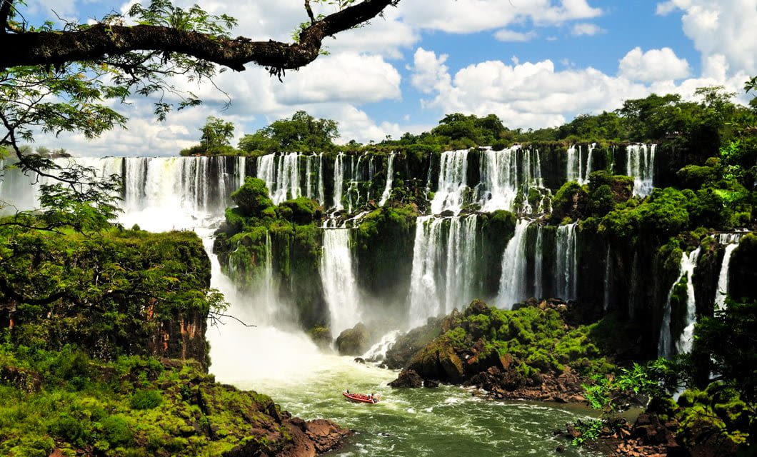 A panoramic view of Iguazu Falls, featured on our Coast to Coast Tour, with multiple waterfalls cascading over lush green cliffs, mist rising from the plunging water, and a small boat visible near the base of the falls.