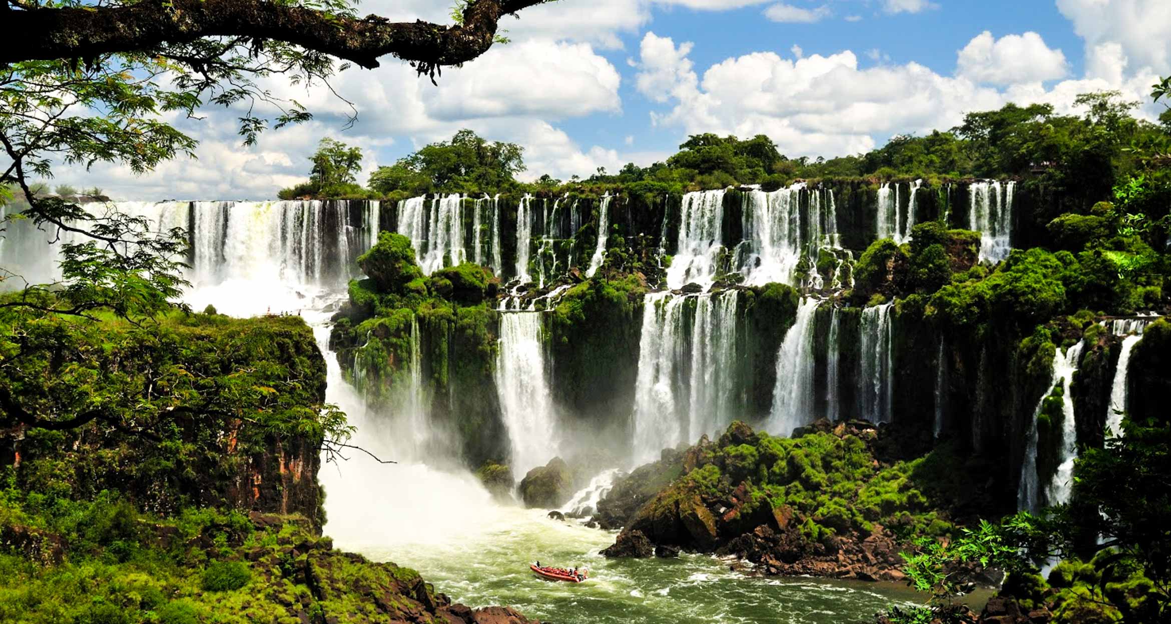 A panoramic view of Iguazu Falls, near the borders of Asuncion and the wild Chaco, shows multiple waterfalls cascading over lush cliffs; a small boat is seen below, surrounded by mist and dense vegetation.