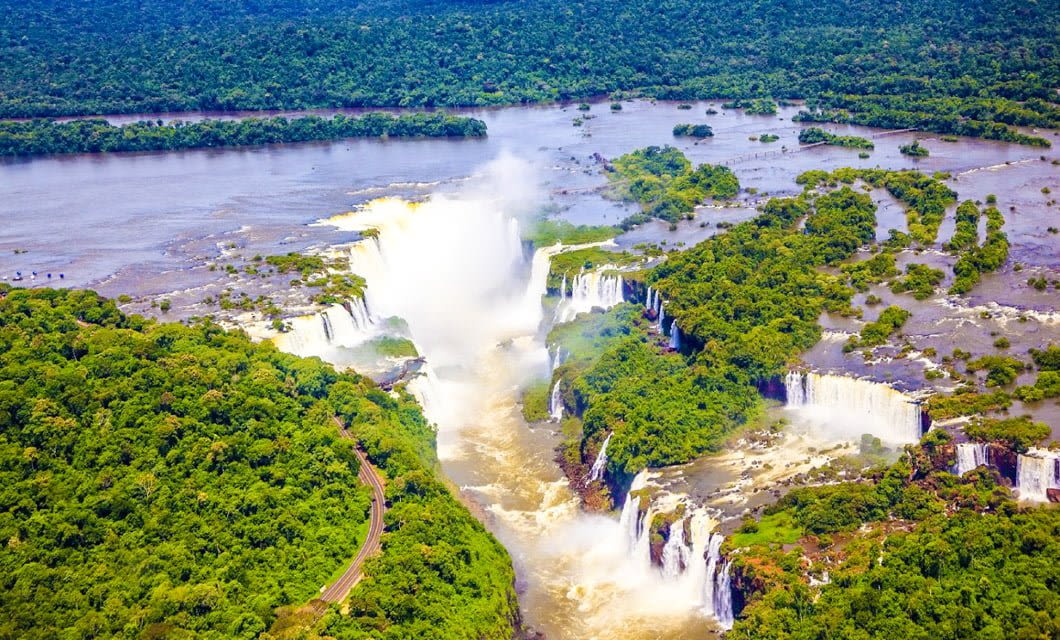 Aerial view of Iguassu Falls surrounded by lush green rainforest, with wide rivers flowing over multiple waterfalls and mist rising above the cascades—a breathtaking scene near destinations known for Brazil farm visits.