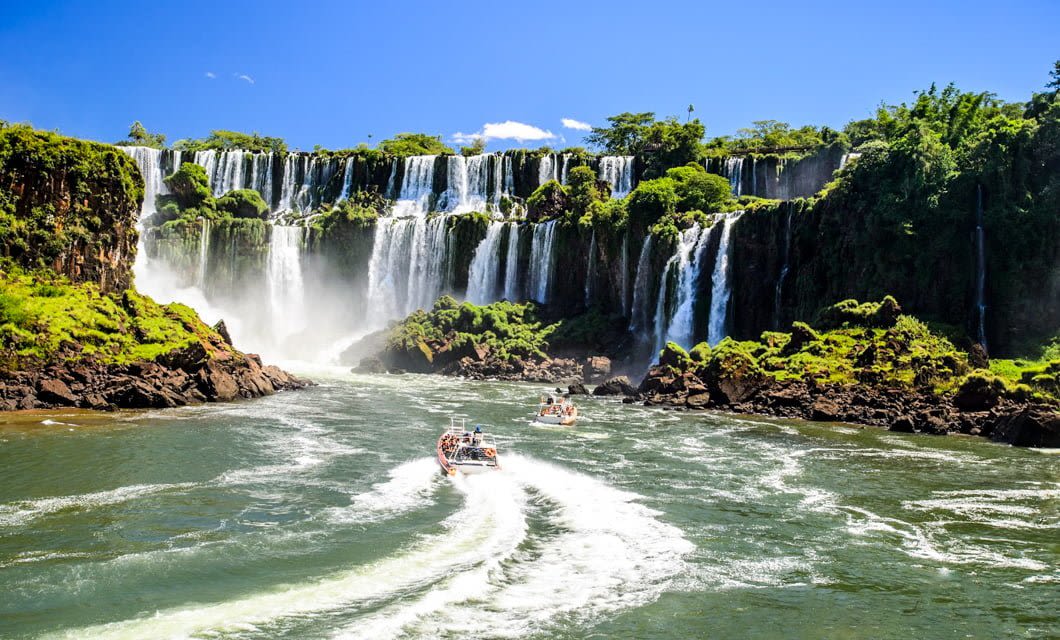 Two boats with tourists approach the base of a large, multi-tiered waterfall surrounded by lush greenery under a bright blue sky—an unforgettable sight on the Historic Rail Tour along the Brazil Gold Route. Turbulent waters and mist add to the adventure.