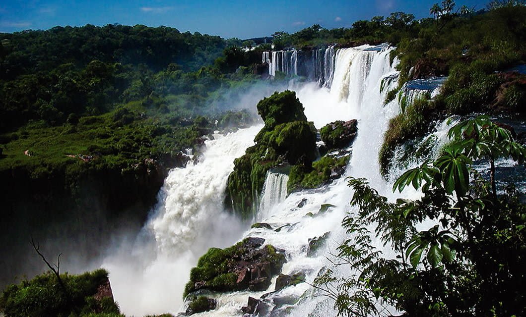 A wide, powerful waterfall cascades over lush, green cliffs surrounded by dense forest under a blue sky, as the mist and white water below seem to dance with the lively rhythm of samba.