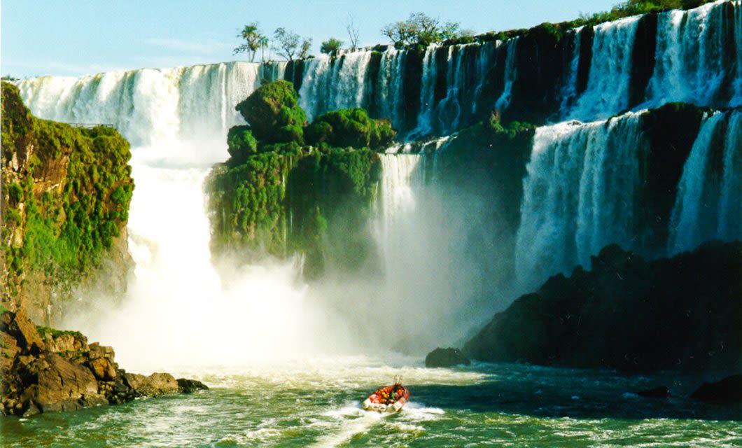 A small boat with people approaches the base of Iguazu’s tall, powerful waterfalls surrounded by lush greenery and rocky cliffs, with mist rising from the cascading water under a clear blue sky—a highlight of any Paraguay tour.