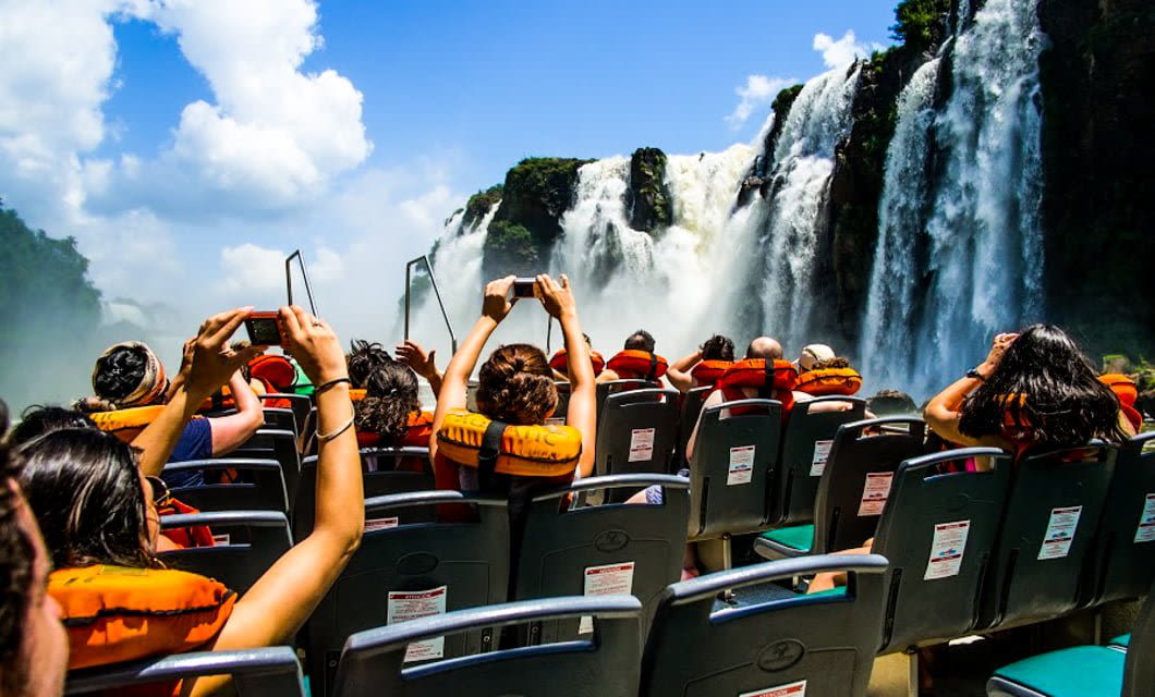 People wearing orange life jackets sit on a boat in Argentina, Land of Contrast, taking photos of large waterfalls cascading over rocks beneath a bright blue sky. The lively scene is filled with excitement and natural beauty.