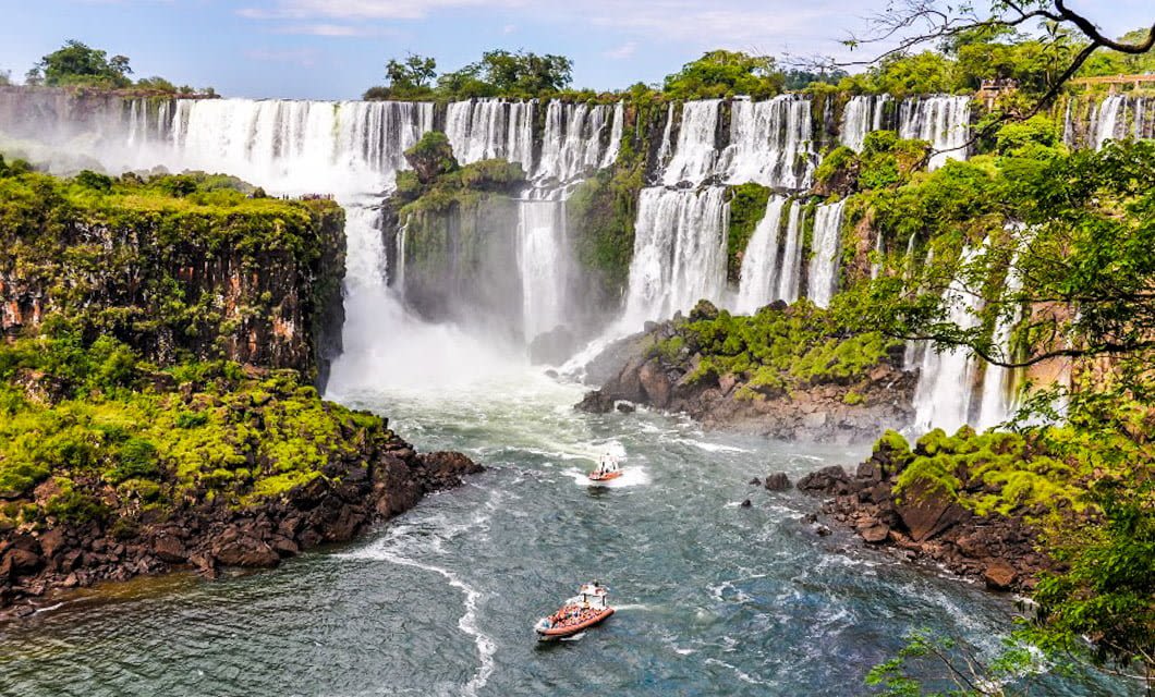 Large waterfalls cascade down lush green cliffs into a wide river below at Iguazú Falls. Several boats with tourists are on the water near the base of the falls, surrounded by dense vegetation, mist, and echoes of ancient Jesuit Missions.