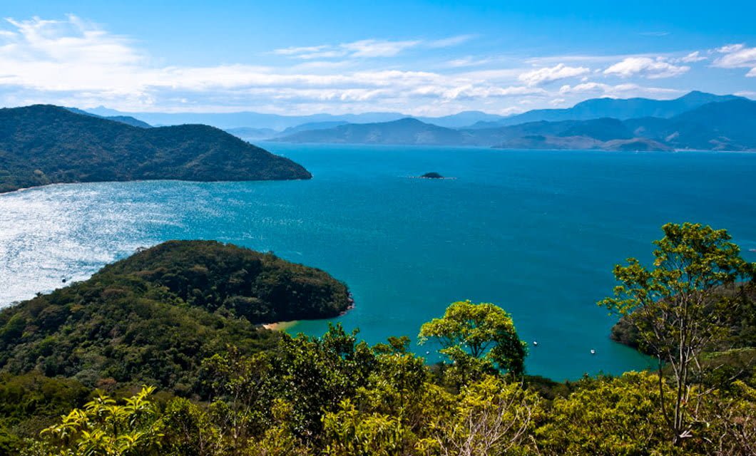 A scenic view of Rio’s blue ocean bay, surrounded by lush green hills and distant mountains, captures the tropical island charm near Paraty under a clear sky with scattered clouds.