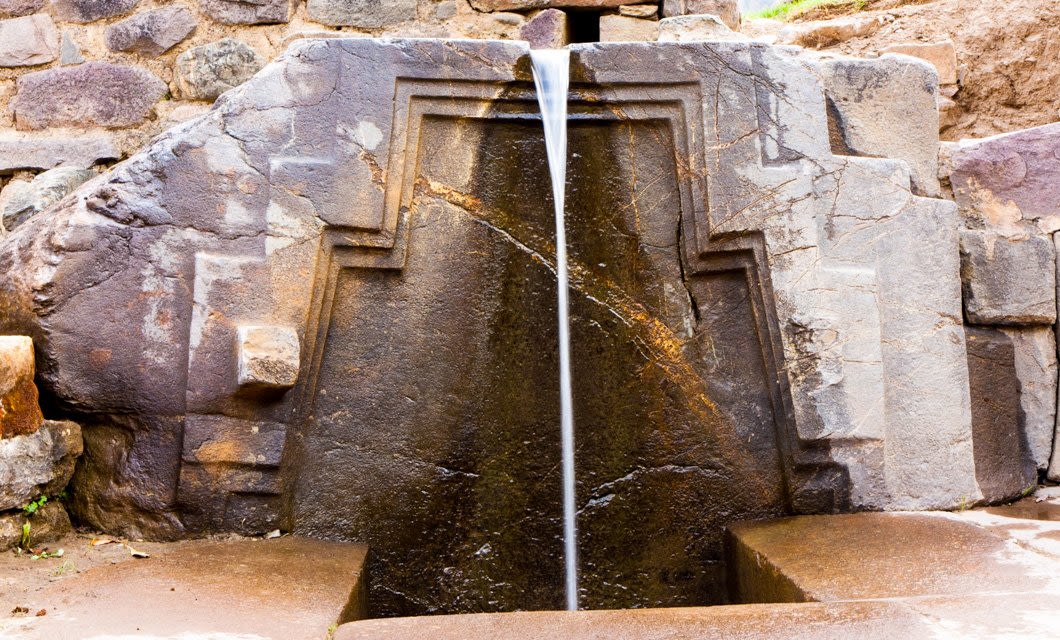 A thin stream of water flows from a stone spout set in an ancient, geometrically-carved stone wall along the Lares Trek, filling a small basin below. The structure features precise lines and shapes.