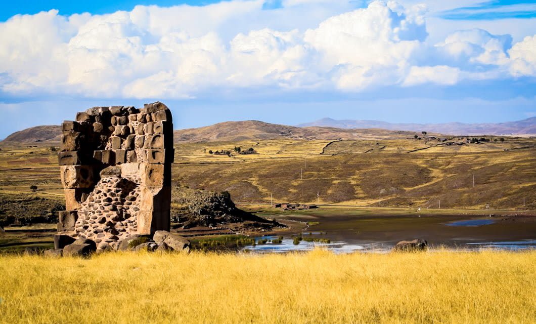 A stone ruin stands alone in a field of tall yellow grass, echoing the essence of Peru, with rolling hills, a small lake, and scattered clouds in the background under a bright blue sky.