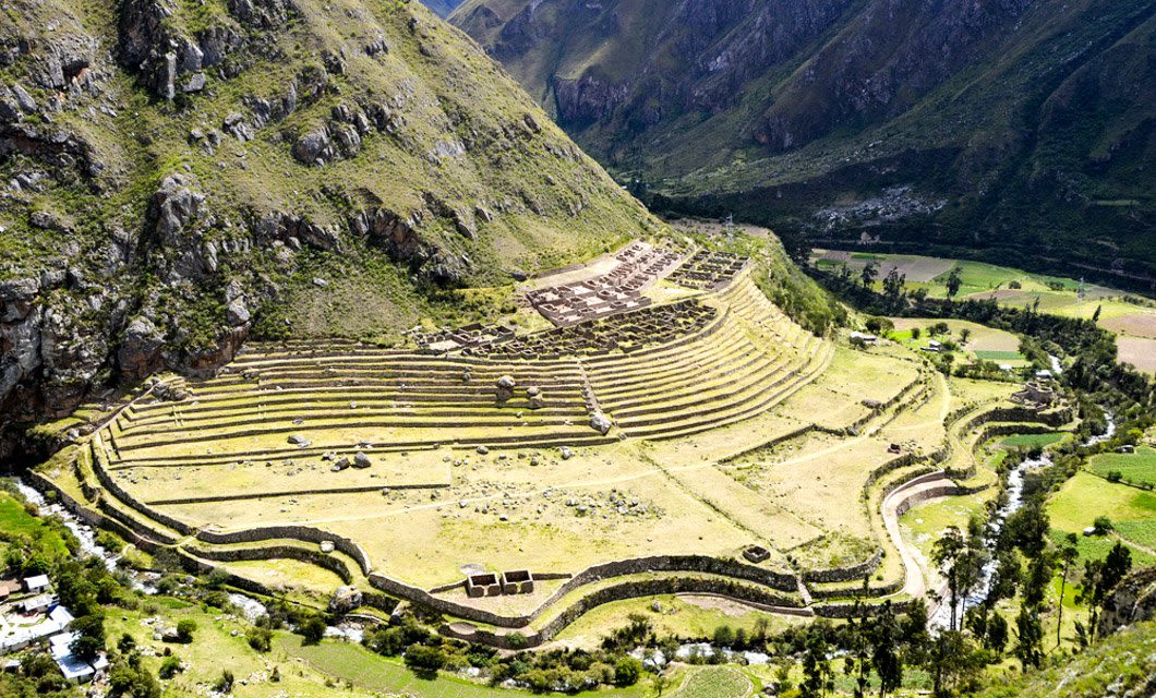 Aerial view of ancient agricultural terraces and ruins built on a sloped mountainside, surrounded by green valleys and rugged peaks, along the scenic Salkantay Trek near Salkantay Lodge in the rural Andean landscape.