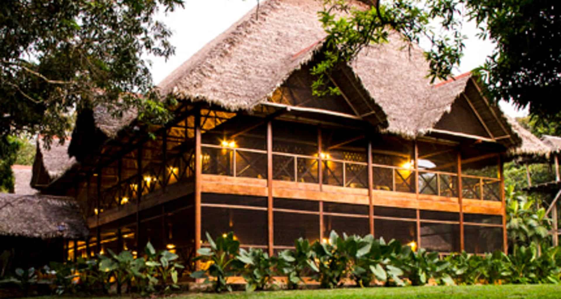 A large two-story Amazon lodge with a thatched roof and wooden frame, warmly lit from within, surrounded by lush green plants and trees—reminiscent of Peru’s Inkaterra Hacienda Concepción.