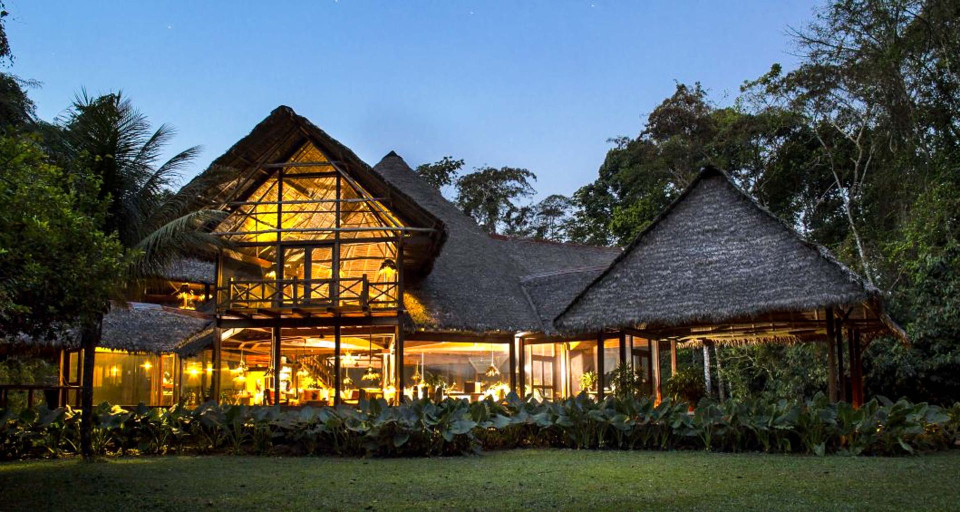 A large Jungle Lodge with thatched roofs is warmly lit at dusk, surrounded by lush greenery and tall trees in the Amazon Rainforest. The glowing interior is visible through large glass windows, evoking the charm of Inkaterra Reserva Amazonica.