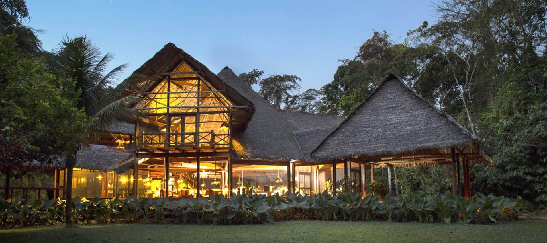 A large thatched-roof lodge with warm lights shining through open walls sits surrounded by lush tropical trees and plants at dusk, offering an inviting retreat for rainforest tours in the heart of the Peruvian Amazon.