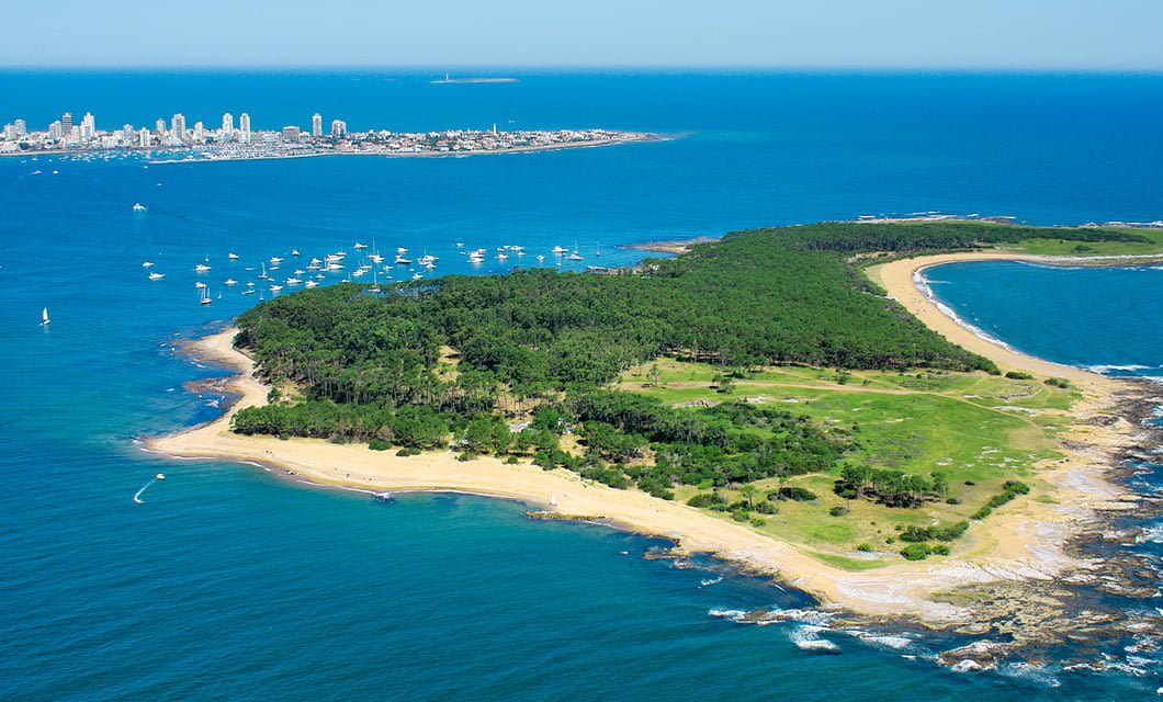 Aerial view of a lush, green island with sandy beaches and rocky shores surrounded by blue ocean. Part of the Coast to Coast Tour, with a city skyline and boats visible in the distance across the water.