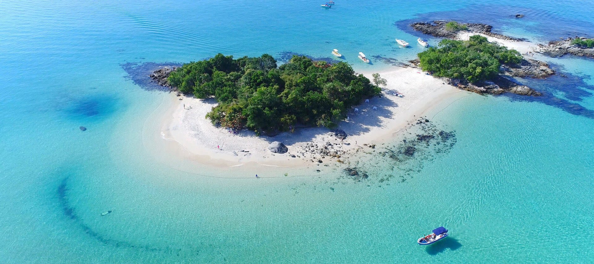 A small, lush island with white sandy beaches is surrounded by clear turquoise water; several boats are anchored near the shore, and a small blue boat is approaching this hidden gem among Brazil’s stunning beach resorts.